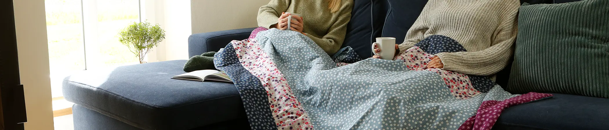 Zwei Personen sitzen in beigen Pullovern auf einem blauen Sofa, zugedeckt mit einer gemusterten Decke, halten je eine Tasse. Neben ihnen liegt ein aufgeschlagenes Buch. Im Hintergrund steht eine Topfpflanze.