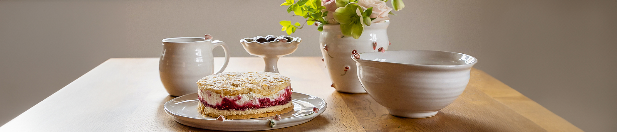 Heller Holztisch mit Sahnetorte, wei&szlig;en Keramikschalen, Wasserkrug und Blumenstrau&szlig;, weiche Farben und zarte Texturen.
