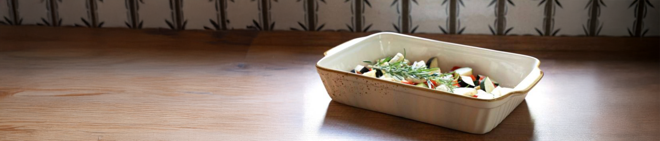 Eine helle Auflaufform gefüllt mit Tomaten, Zucchini und Aubergine steht auf einer Holz-Arbeitsplatte vor einer gefliesten Wand