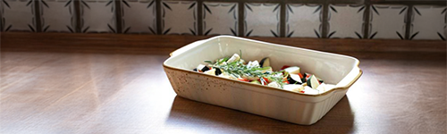 Eine helle Auflaufform gefüllt mit Tomaten, Zucchini und Aubergine steht auf einer Holz-Arbeitsplatte vor einer gefliesten Wand