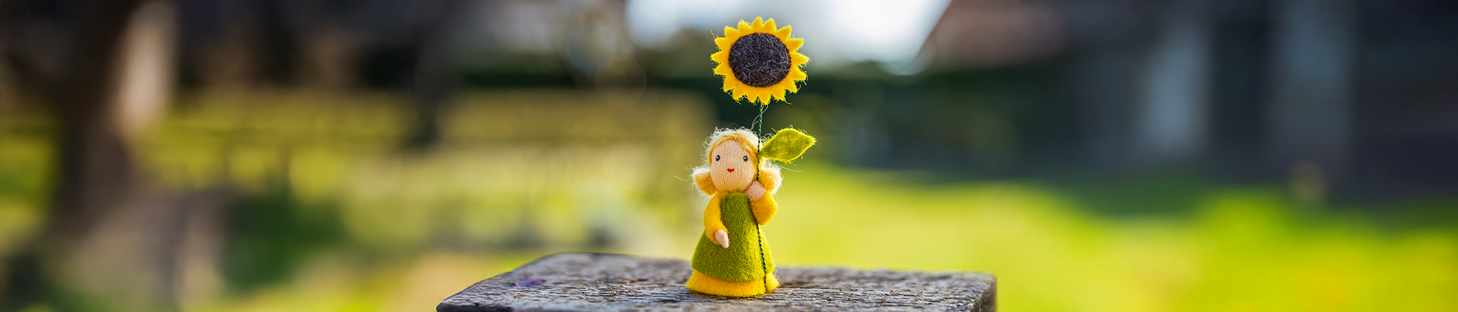 Headerbild Neuheiten zeigt gelbe Filzpuppe mit gr&uuml;ner Kleidung und Sonnenblume, unscharfer Hintergrund mit leuchtendem Gr&uuml;n und Naturtextur.
