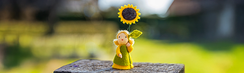 Headerbild Neuheiten zeigt gelbe Filzpuppe mit gr&uuml;ner Kleidung und Sonnenblume, unscharfer Hintergrund mit leuchtendem Gr&uuml;n und Naturtextur.