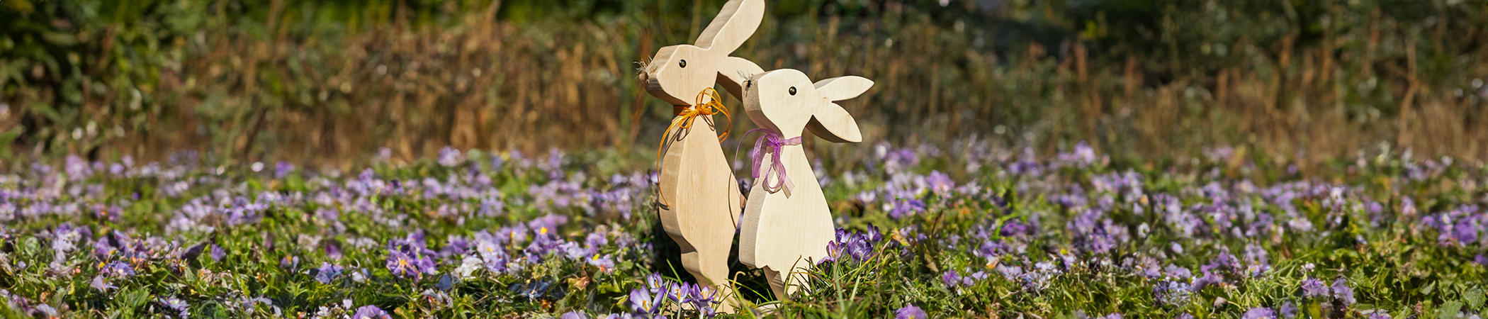 Zwei helle Holzhasen mit Schleifen stehen auf einer Wiese mit lila und gr&uuml;nen Fr&uuml;hlingsblumen.