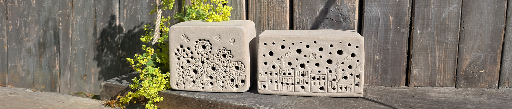 Zwei hellgraue rechteckige Nisthilfen mit dekorativen Mustern und L&ouml;chern stehen auf h&ouml;lzerner Bank neben gr&uuml;nem Blattwerk.