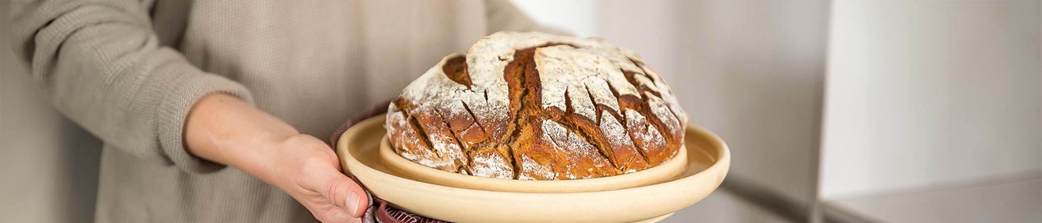 Ein frisches rundes Brot liegt in der Mitte einer cremefarbenen Keramik-Brotbackplatte. Eine Person im beigen Oberteil hält die Platte in der Hand.