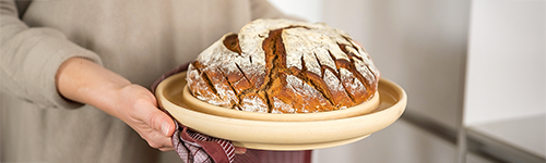 Ein frisches rundes Brot liegt in der Mitte einer cremefarbenen Keramik-Brotbackplatte. Eine Person im beigen Oberteil hält die Platte in der Hand.