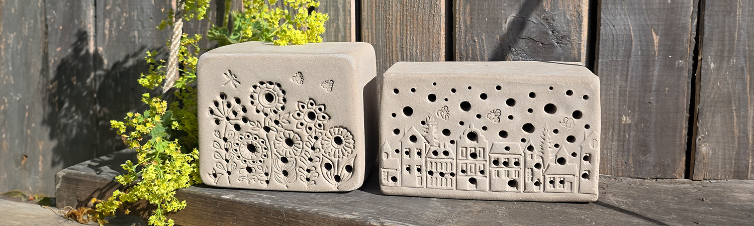 Zwei hellgraue rechteckige Nisthilfen mit dekorativen Mustern und L&ouml;chern stehen auf h&ouml;lzerner Bank neben gr&uuml;nem Blattwerk.