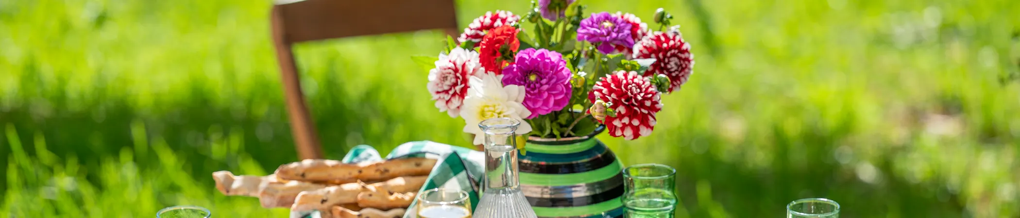 Bunter Blumenstrauß in einer gestreiften Vase auf einem Gartentisch. Daneben eine Glaskaraffe mit Wasser, zwei Gläser und ein Korb mit Brotstangen. Der Hintergrund zeigt unscharfe grüne Wiesen und einen Holzstuhl.