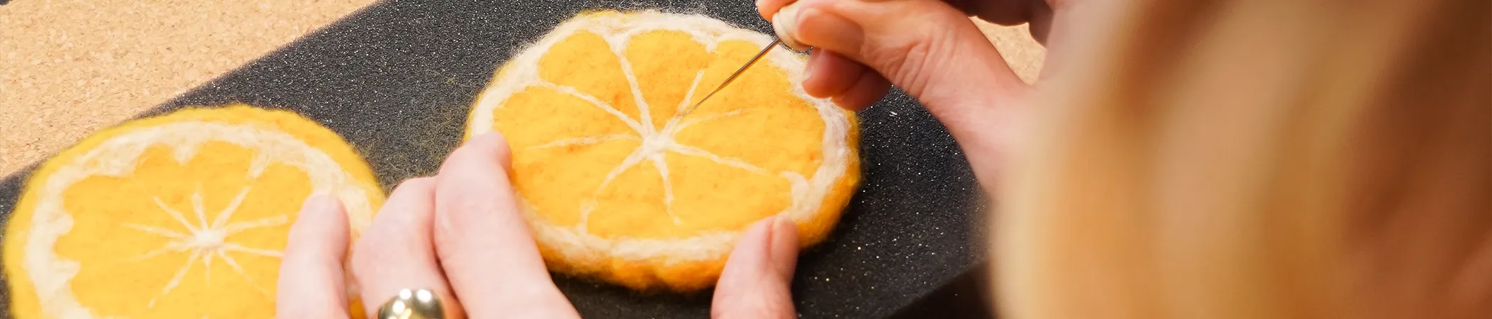 Hände einer Person filzen eine orangefarbene Filzscheibe mit einer Nadel auf schwarzem Hintergrund. Die Filzscheibe ist kreisförmig und ähnelt einer Orangenscheibe mit weißen Segmentlinien.
