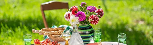 Bunter Blumenstrauß in einer gestreiften Vase auf einem Gartentisch. Daneben eine Glaskaraffe mit Wasser, zwei Gläser und ein Korb mit Brotstangen. Der Hintergrund zeigt unscharfe grüne Wiesen und einen Holzstuhl.
