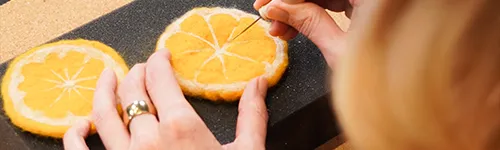 Hände einer Person filzen eine orangefarbene Filzscheibe mit einer Nadel auf schwarzem Hintergrund. Die Filzscheibe ist kreisförmig und ähnelt einer Orangenscheibe mit weißen Segmentlinien.
