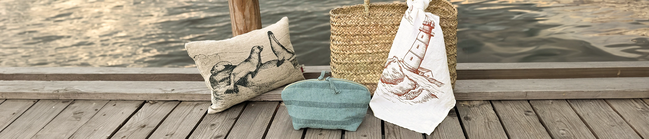 Kissen mit Robbenmotiv, blaues Necessaire, Korbtasche mit Leuchtturmmotivtuch auf Holzsteg am Wasser platziert.