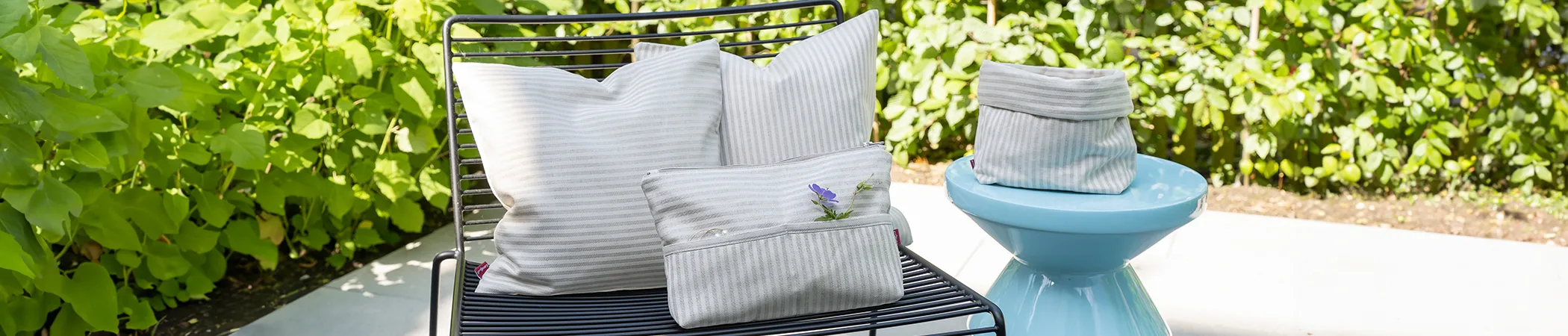 Kissen, Kosmetiktasche und Körbchen aus gestreiftem Landlust-Jubiläumsstoff platziert auf einem Stuhl und einem Hocker auf einer Terrasse in einem Garten