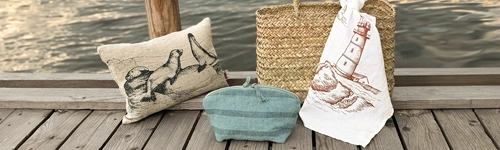 Kissen mit Robbenmotiv, blaues Necessaire, Korbtasche mit Leuchtturmmotivtuch auf Holzsteg am Wasser platziert.