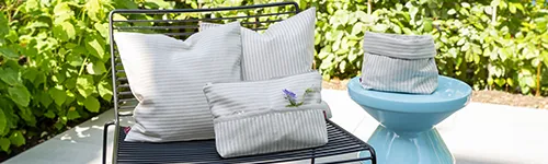 Kissen, Kosmetiktasche und Körbchen aus gestreiftem Landlust-Jubiläumsstoff platziert auf einem Stuhl und einem Hocker auf einer Terrasse in einem Garten