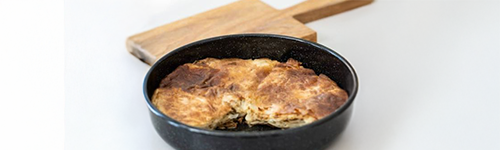 Eine Form aus Emaille in der Kartoffelgratin zubereitet wurde. Im Hintergrund liegt ein Holzbrett