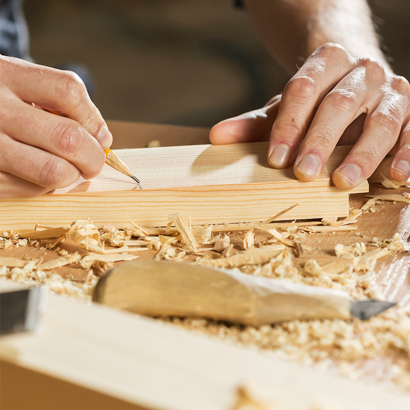 Eine Person zeichnet mit Bleistift etwas auf ein Holzstück. Drum herum liegen Späne