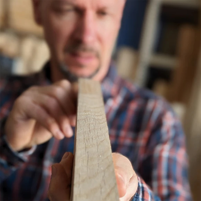 Ein Mann im blau-rot karierten Hemd hält ein Stück Holz in der Hand und schaut es sich ganz genau an