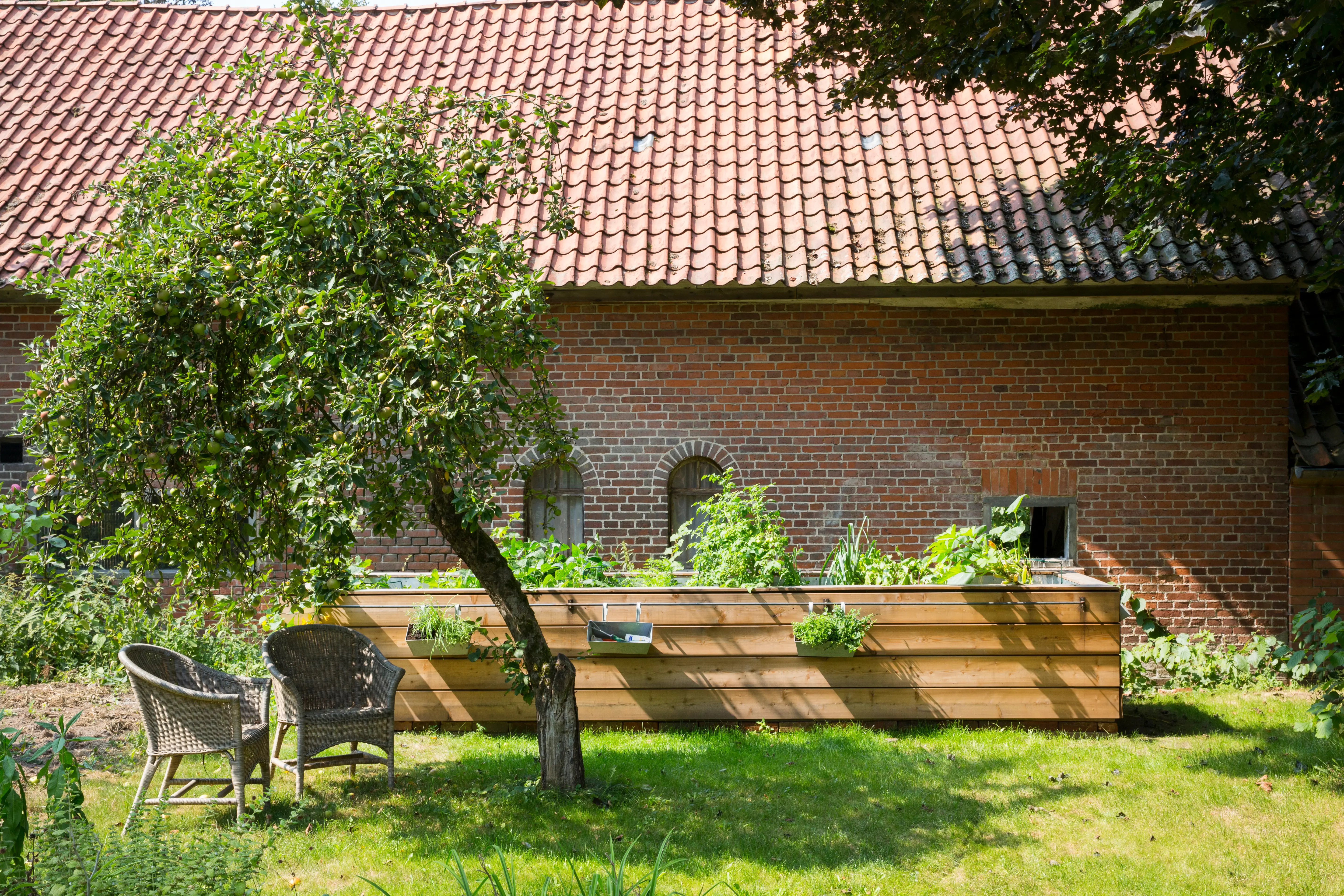 Ein langes Hochbeet mit Holzplanken Design, steht in einem Garten. Vor ihm steht ein Baum und zwei Gartenst&uuml;hle.
