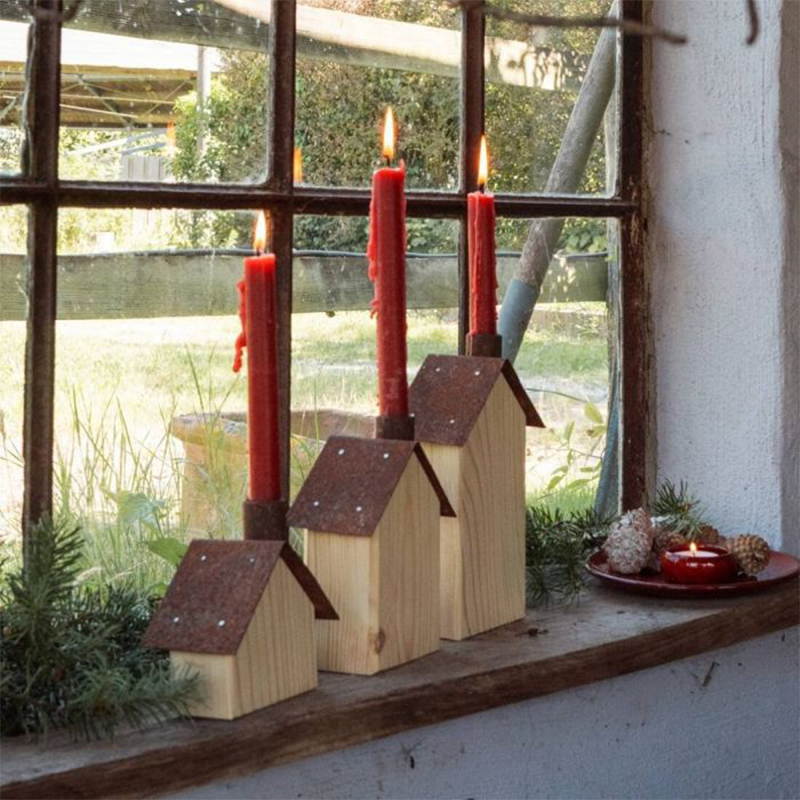 Auf einer Fensterbank stehen drei Holzhäuser mit rostfarbenden Dächern. In den Schornsteinen stecken rote Stabkerzen