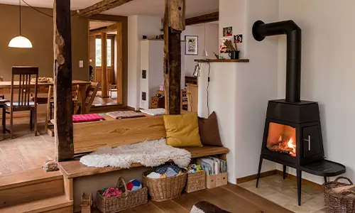 Gemütliches Wohnzimmer mit einem modernen Kaminofen an der rechten Wand, Sitzgelegenheiten aus Holz mit Kissen in der Mitte, Bücher und Körbe darunter, und ein Essbereich im Hintergrund links. Warme Holzakzente dominieren den Raum. Bild verlinkt zur Seite