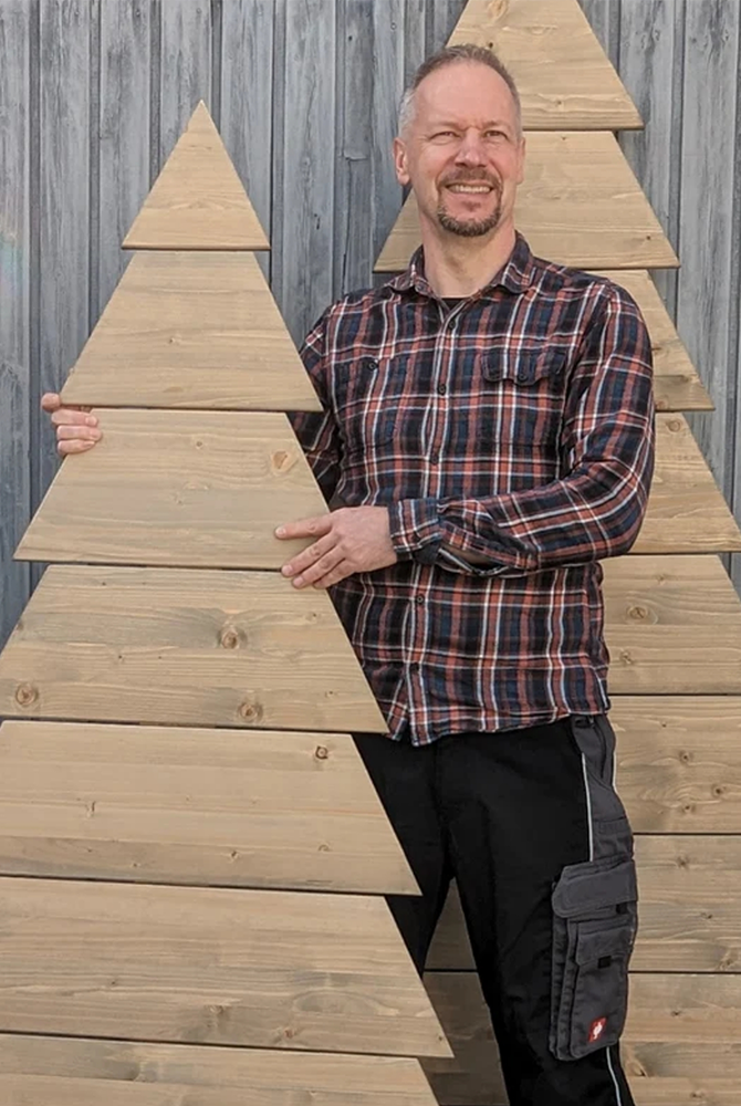 Ein Mann steht mit Holzobjekten in Form von Tannenbäumen vor einer Holzwand