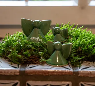 Drei grüne Keramikfiguren in Form von Engeln stehen auf einer Fensterbank umgeben von Moos.