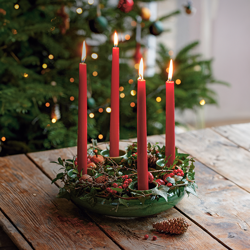 Ein Kranz aus grünem Keramik auf dem vier rote Stabkerzen brennen. Er ist mit Tannenzweigen und Beeren geschmückt. Im Hintergrund ist ein Weihnachtsbaum zu erkennen.