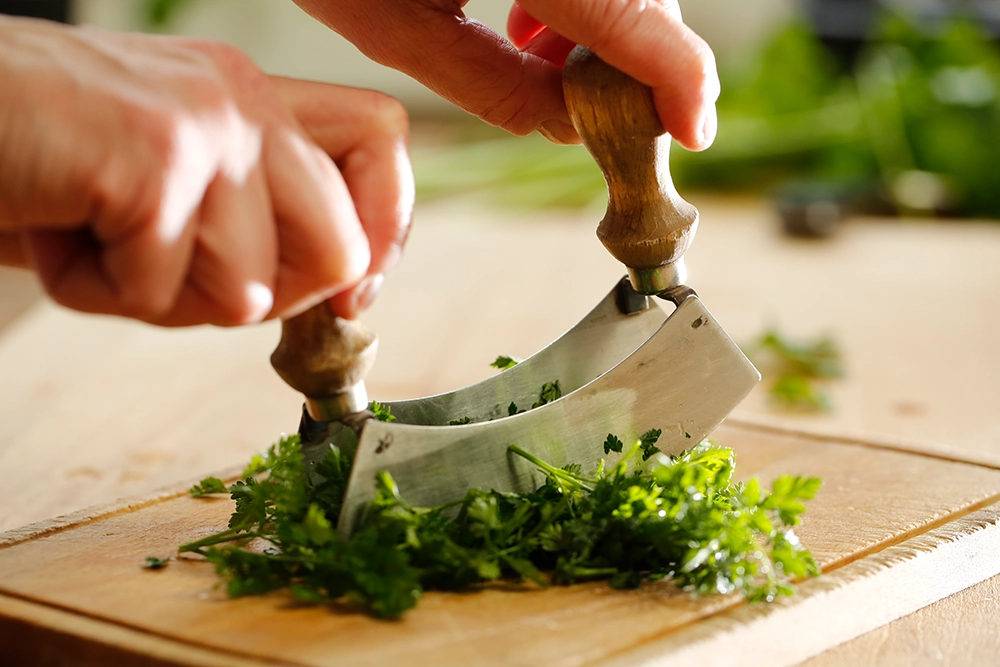 Zwei Hände halten ein Wiegemesser und schneiden frische grüne Kräuter auf einem Holzbrett. Im Hintergrund sind unscharf weitere Kräuter zu sehen.Bild verlinkt zum Rezept