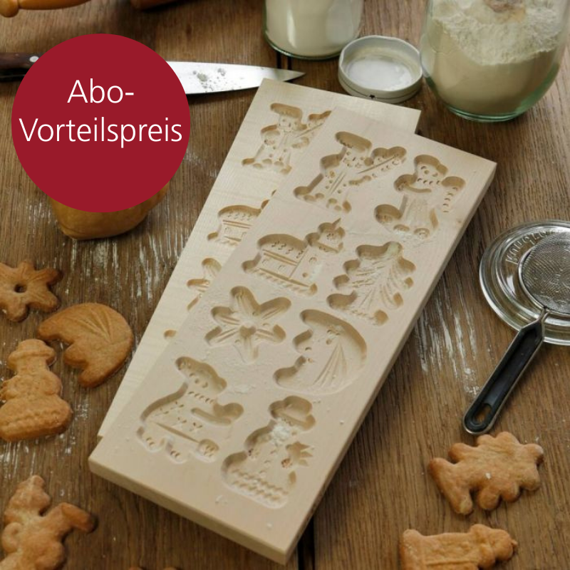 Eine Holzform in die acht weihnachtliche Motive reingeritzt worden sind. Auf der Holztischplatte darunter liegen weitere Backutensilien, Keksteig und Spekulatiuskekse.