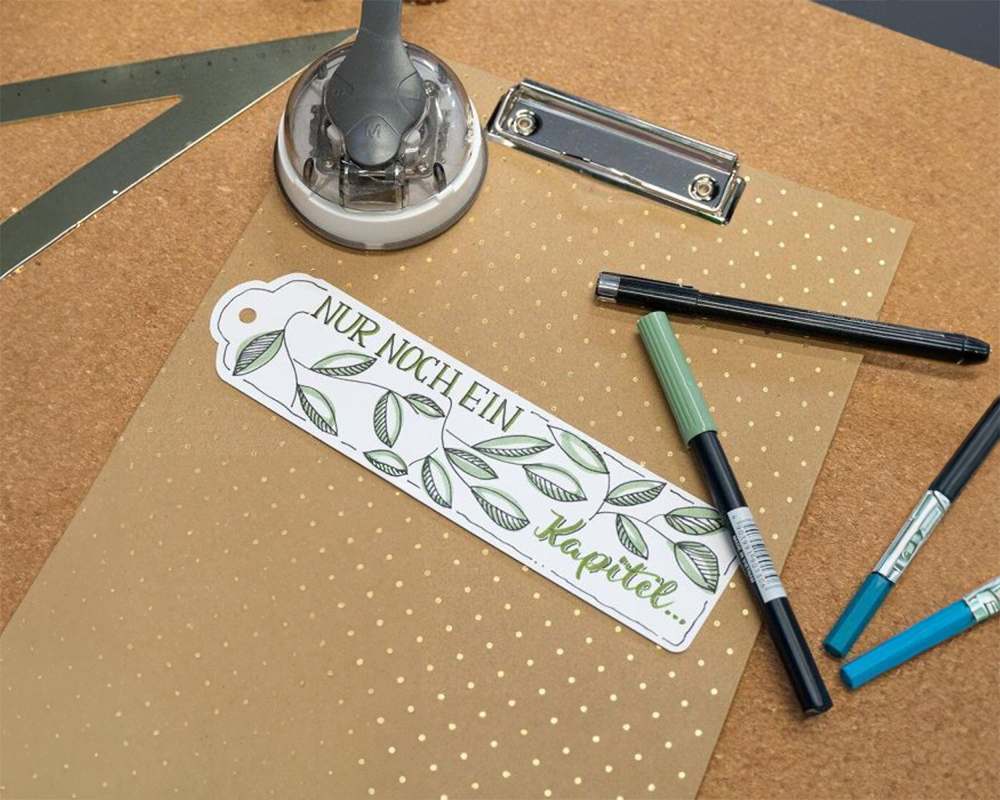 Auf braunem Papier mit goldenen Punkten liegt ein weißes Lesezeichen, dass mit schöner Schrift und Blättern verziehrt ist. Darauf steht "Nur noch ein Kapitel...". Drumherum liegen verschiedene Stifte und andere Utensilien