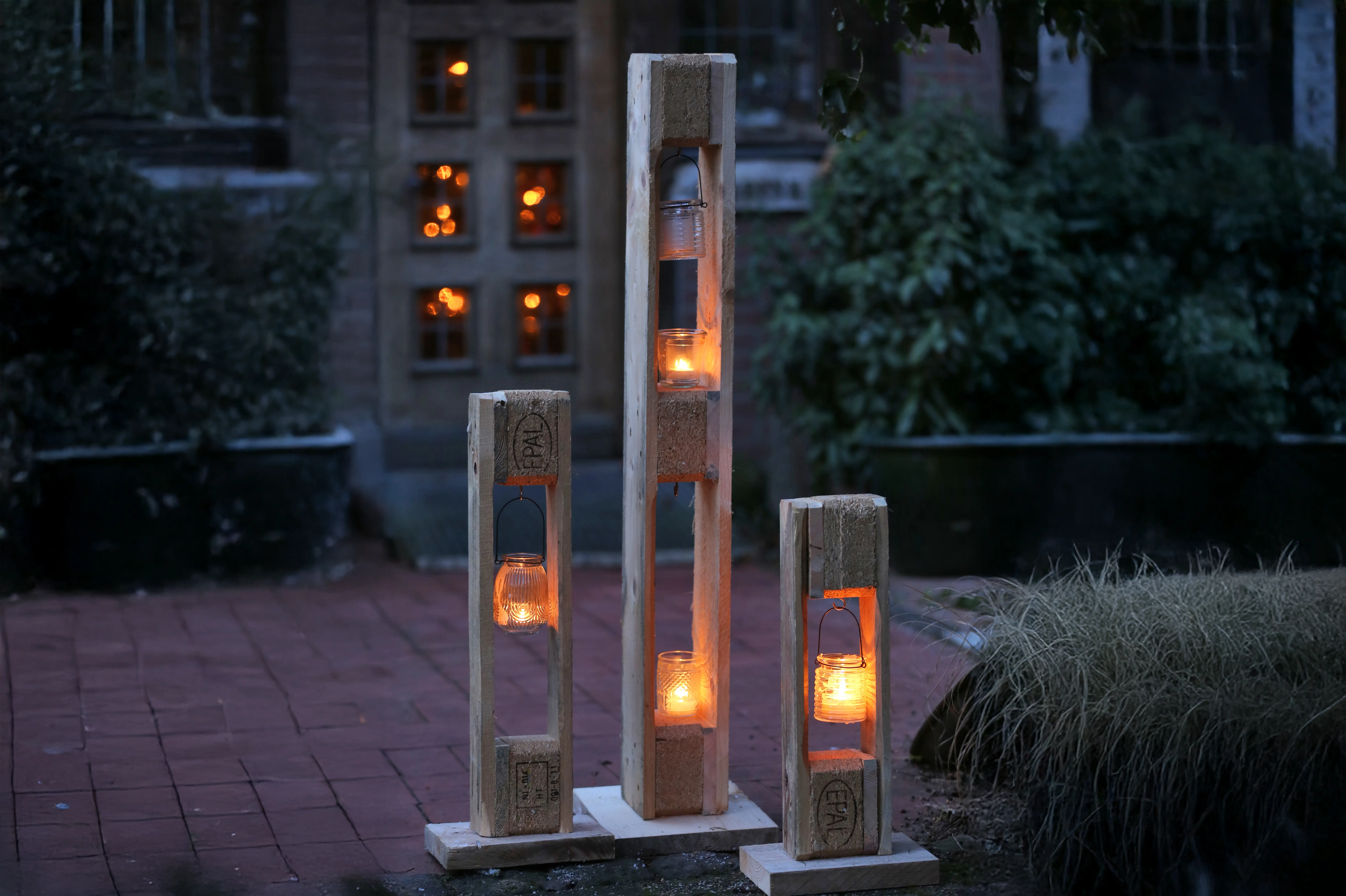S&auml;ulen aus recycleten Holzpaletten stehen im dunkeln vor einem Hauseingang. Innerhalb der Palleten sind kleine Windlichter angebracht.