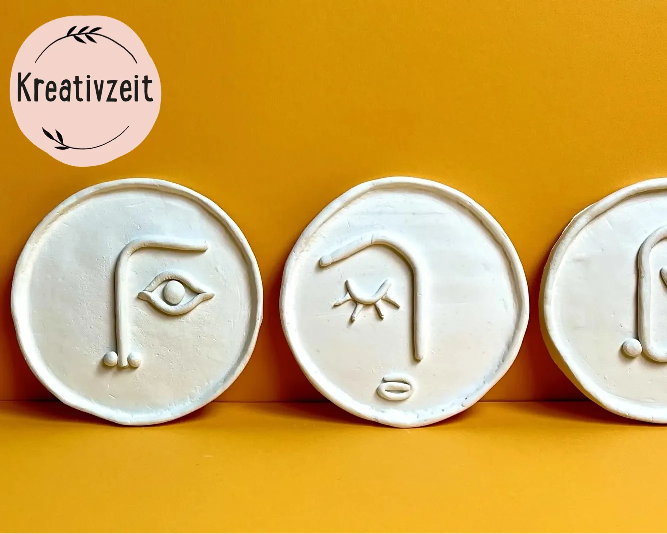 Drei runde Tonplatten mit abstrakten Gesichtern, nebeneinander auf einem orangenen Hintergrund platziert. Links ein Logo mit stilisierten Blättern und dem Wort Kreativzeit. Bild verlinkt zum Workshop.