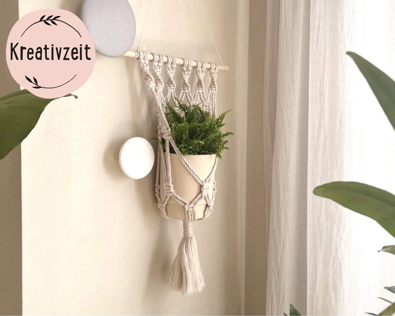 Wanddekoration mit Makramee-Pflanzenhalter, der einen topfgebundenen Farn enthält. Im Hintergrund helle Vorhänge, Pflanzenblätter im Vordergrund. Oben links brauner Kreis mit der Aufschrift "Kreativzeit", Bild verlinkt zum Workshop.