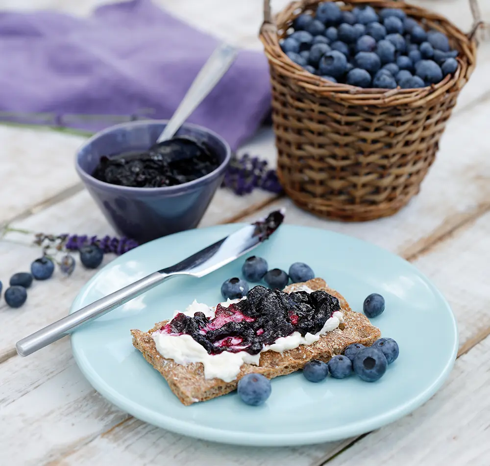 Ein Knäckebrot auf einem hellblauem Teller, das mit Frischkäse und dunkler Marmelade beschmiert ist. Drum herum liegen verstreut ein paar Blaubeeren. Im Hintergrund ist eine Schüssel mit mehr Marmelade und ein Korb mit Blaubeeren zu sehen.