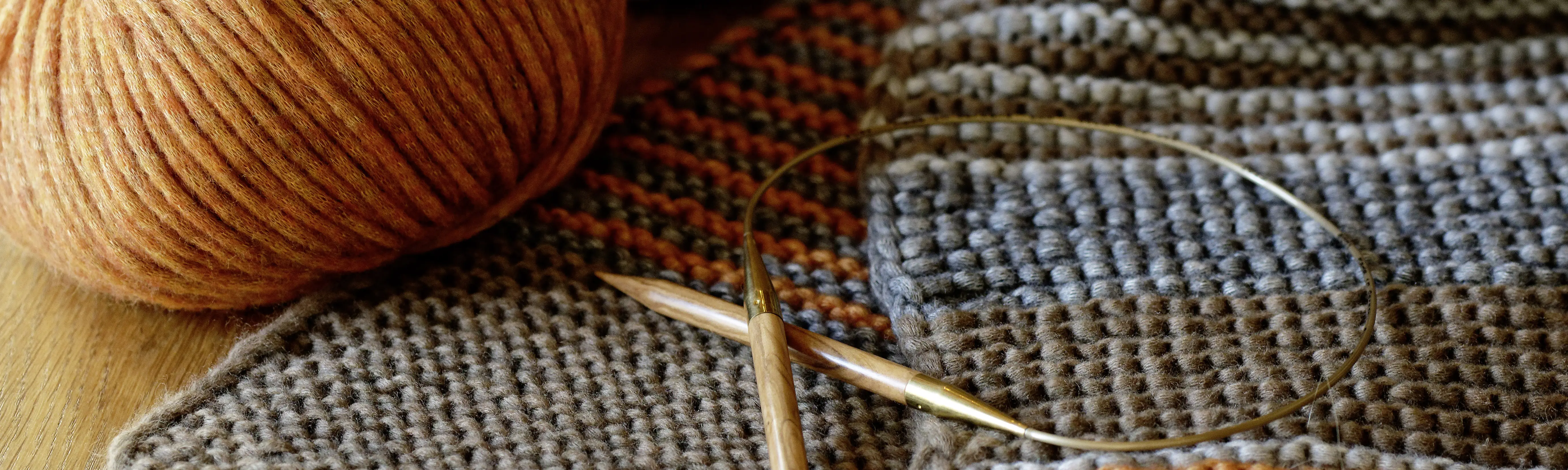 Eine Stricknadel aus Holz liegt neben einem orange-braunen Wollkn&auml;ul auf einem gestrickten Schal.
