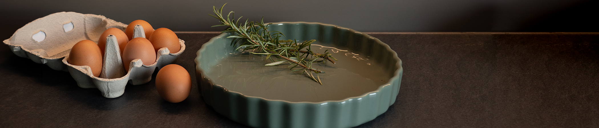 Headerbild Neuheiten zeigt eine Tarteform mit Rosmarinzweigen, umgeben von einem Eierkarton und Arbeitsfläche einer Küche im Hintergrund.