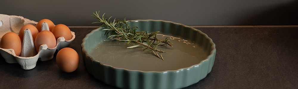 Headerbild Neuheiten zeigt eine Tarteform mit Rosmarinzweigen, umgeben von einem Eierkarton und Arbeitsfläche einer Küche im Hintergrund.