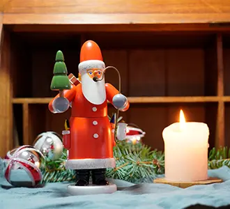 Eine Räuchermännchen-Figur mit dem Aussehen eines Weihnachtsmannes steht auf einem Tisch. Daneben ist eine brennende Kerze, Weihnachtsschmuck und Tannenzweige dekoriert.