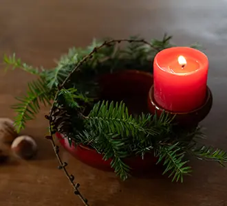 Ein Kranz aus rotem Keramik, auf dem eine rote Kerze steht. Der restliche Kranz ist mit Tannenzweigen und Nüssen dekoriert.
