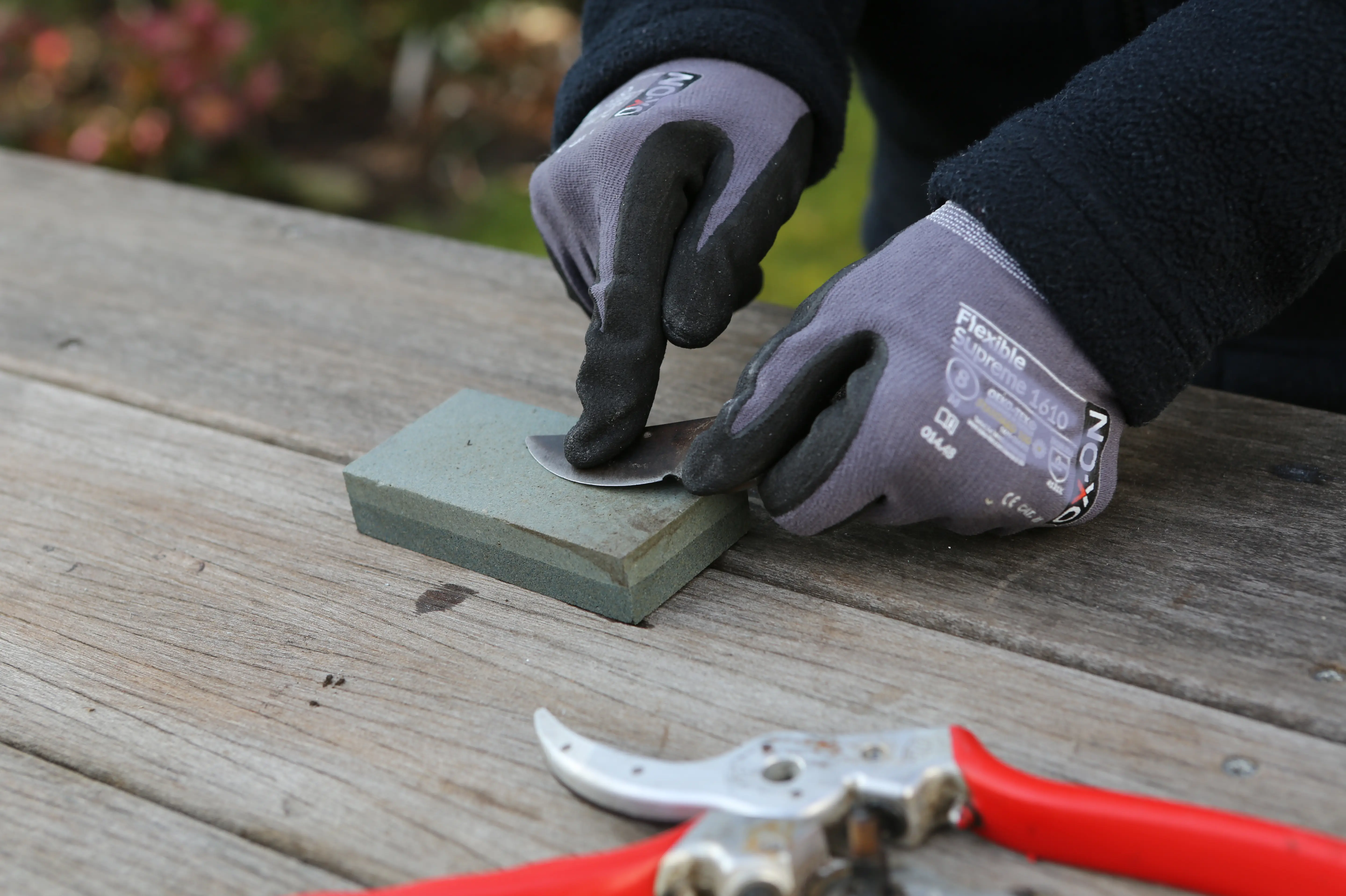 Eine Klinge einer Gartenschere wird von jemandem auf einem Schleifstein mit Handschuhen geschliffen. Die auseinandergebaute Schere liegt daneben auf dem Holztisch.