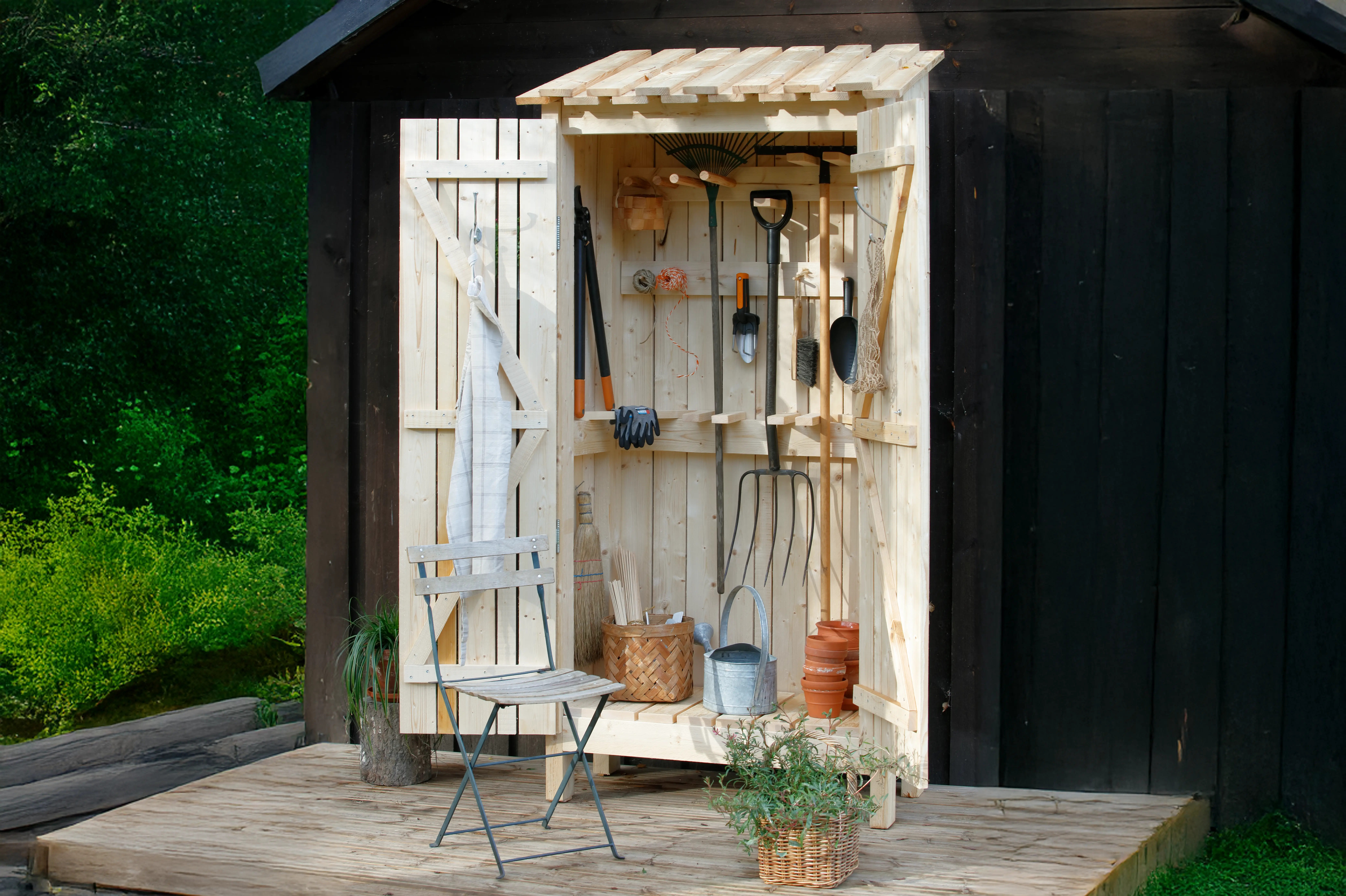 Ein offener Geräte-Holzschrank steht auf einer kleinen Holzerhöhnung, vor einem Haus. Vor dem Schrank steht ein kleiner Gartenstuhl.