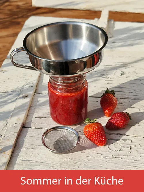 Ein Trichter, der auf einem vollen Marmeladenglas steht. Daneben liegt das zugehörige Sieb und frische Erdbeeren auf einer weißen Holztischplatte. Bild verlinkt zur Landingpage "Sommer in der Küche".