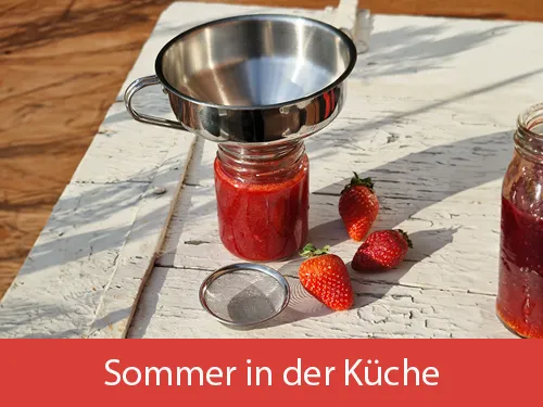 Ein Trichter, der auf einem vollen Marmeladenglas steht. Daneben liegt das zugehörige Sieb und frische Erdbeeren auf einer weißen Holztischplatte. Bild verlinkt zur Landingpage "Sommer in der Küche".