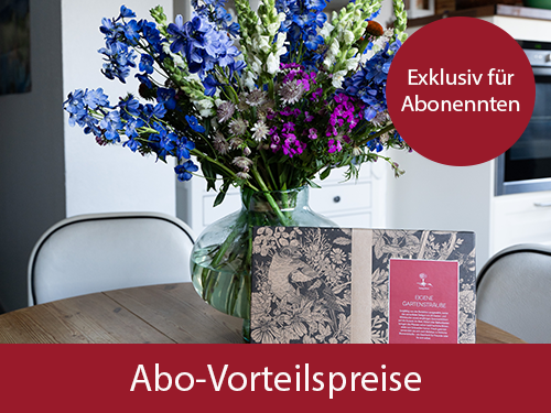 Ein Blumenstrau&szlig; steht auf einem Holztisch. Im Vordergrund befinden sich ein Pappkarton und kleine Papiert&uuml;ten, im Hintergrund ist eine K&uuml;che zu erkennen.