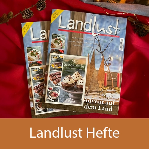 Eine Gruppe von „Landlust“-Magazinen liegt auf einem roten Tuch, umgeben von Tannenzapfen und Zweigen. Ein Heft zeigt Holzbäume auf dem Cover, ein weiteres Heft thematisiert Schokolade. Bild verlinkt zu den Landlust Heften.