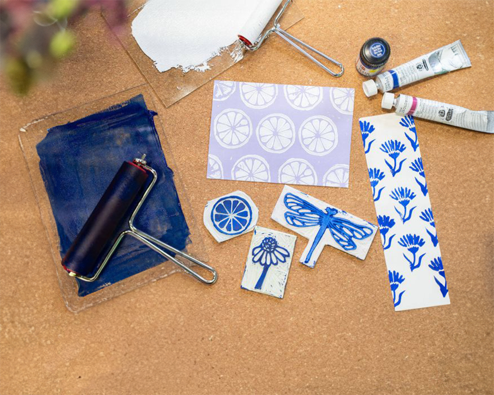 Auf einer braunen Unterlage liegen alle notwendigen Materialien zum Stempeln: Papier, Stempel mit blauer Farbe, Farbbehälter und Rolle