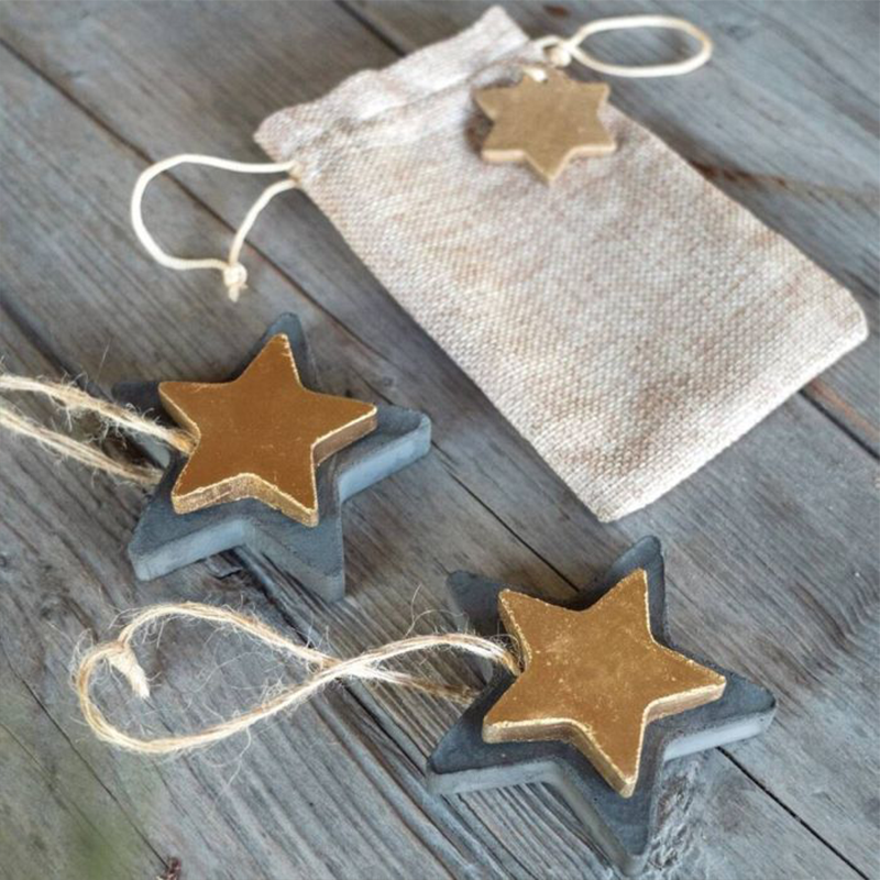 Zwei unterschiedlich große Sterne in gold und anthrazit sind aufeinander geklebt und mit einem Band als Anhänger fertiggestellt. Sie liegen mit einem Jutesäckchen auf einem Holztisch.