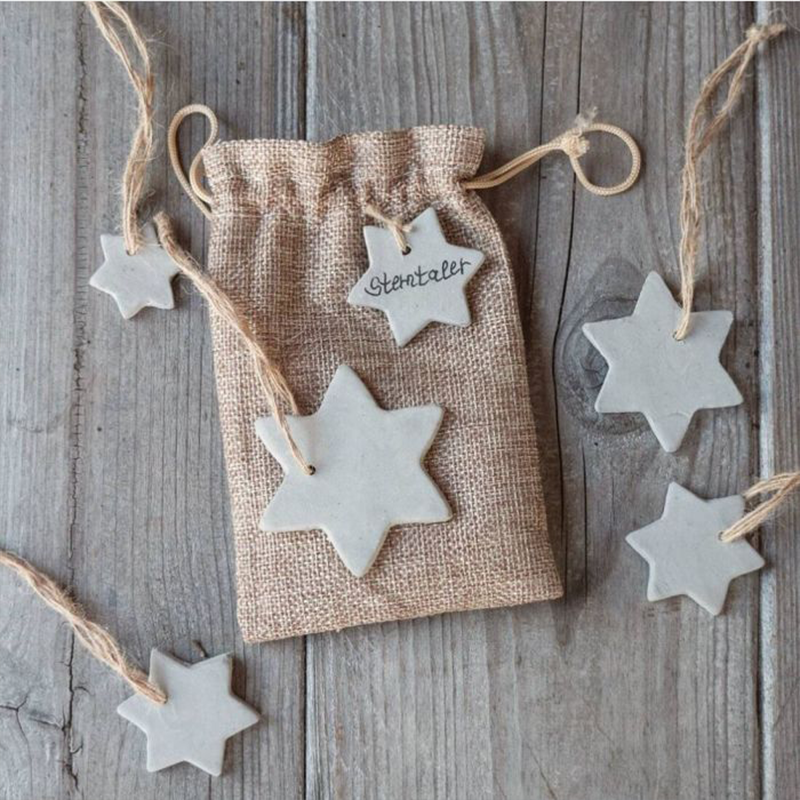 Kleine weiße Anhänger in Sternform liegen mit einem Säckchen aus Jute auf einem Holztisch.