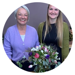 Zwei Frauen stehen l&auml;chelnd vor einem sch&ouml;nen Blumenstrau&szlig; in einer Glasvase.